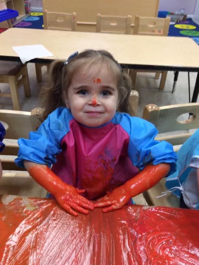 A toddler with paint-covered arms smiling at a messy art table